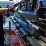 Worker adjusts a large metal sheet on an industrial press, ensuring precision in a busy manufacturing environment.